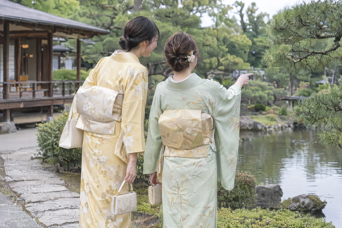 Two friends in pale yellow and pale green houmongi walking by a pond in a Japanese garden, seen from behind