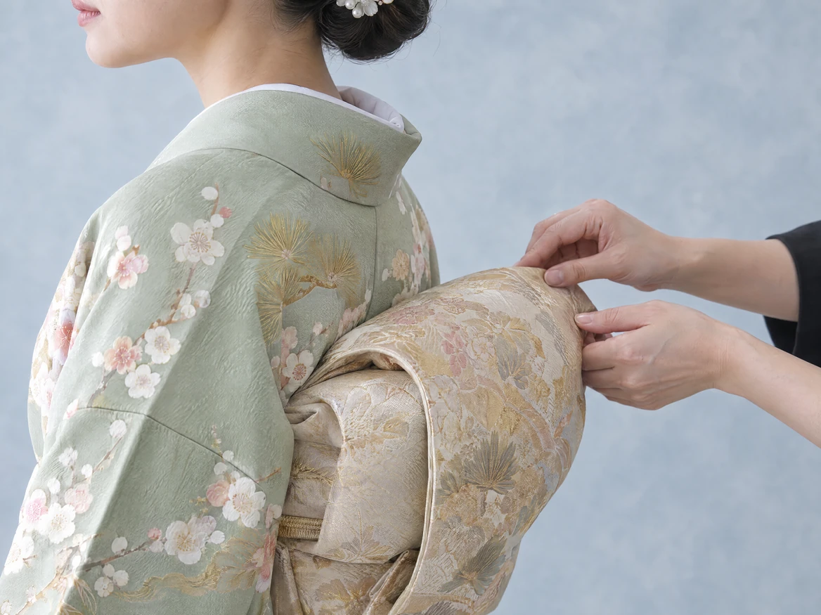 A dresser adjusting the double drum obi knot behind a customer wearing a pale green houmongi