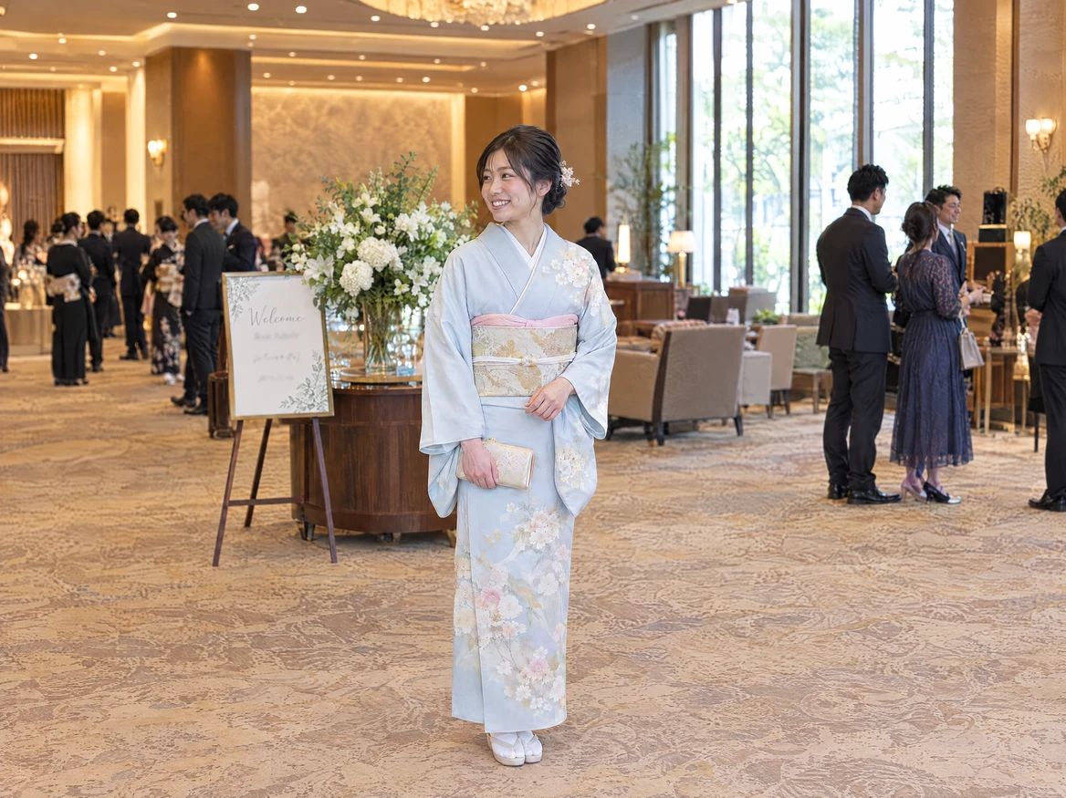 A woman in a light blue houmongi with a gold fukuro obi standing in a hotel lobby before a wedding reception