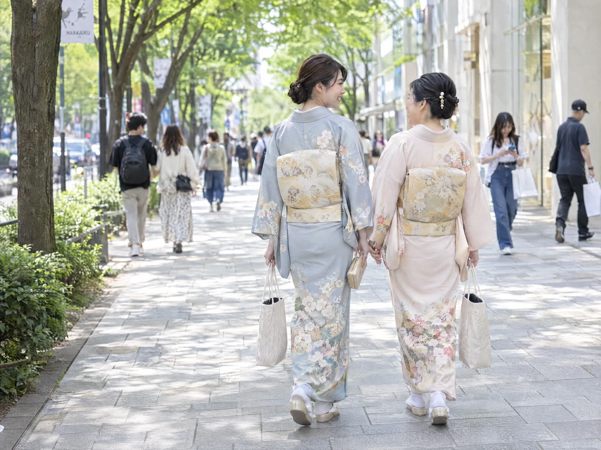 Two friends in light blue and pale pink houmongi chatting as they walk down a tree-lined street in Harajuku, carrying only a kimono handbag and a white sub-bag